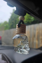 Clear glass skull diffuser hanging from a car's rearview mirror with a blurred outdoor background